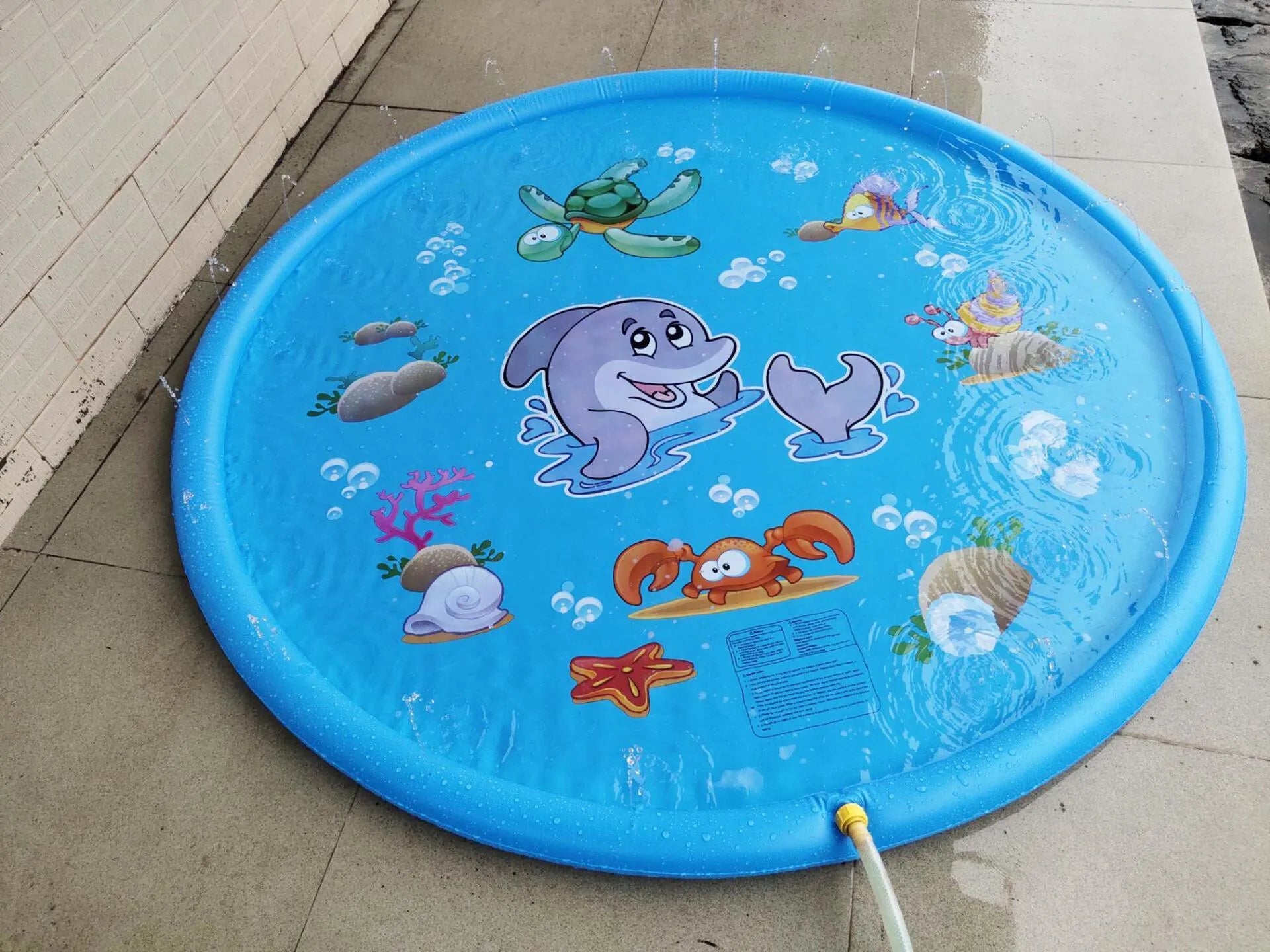 Blue PVC children's water spray mat with marine animal designs laid on grass, connected to a garden hose.