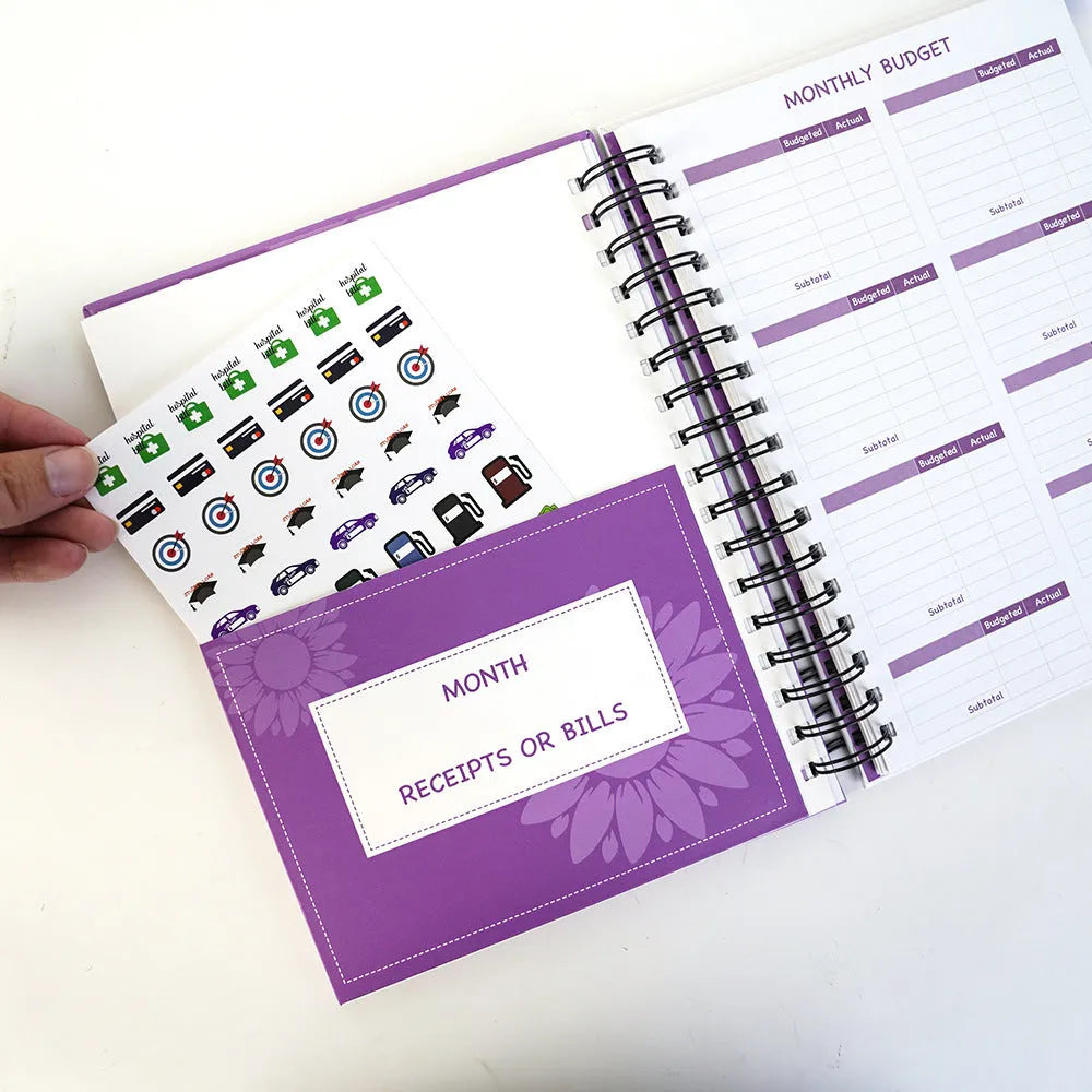 Purple coil-bound budget book open showing monthly financial planning layout with stickers and paper clips