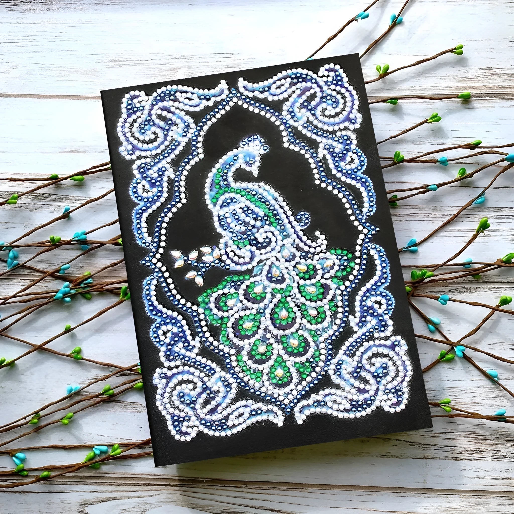 Black A5 notebook with a peacock design made from shaped and bright diamond painting drills.