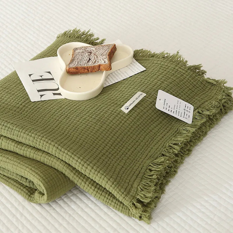 Four-layer gauze cotton blanket with tassel trim in avocado green colour draped over a sofa