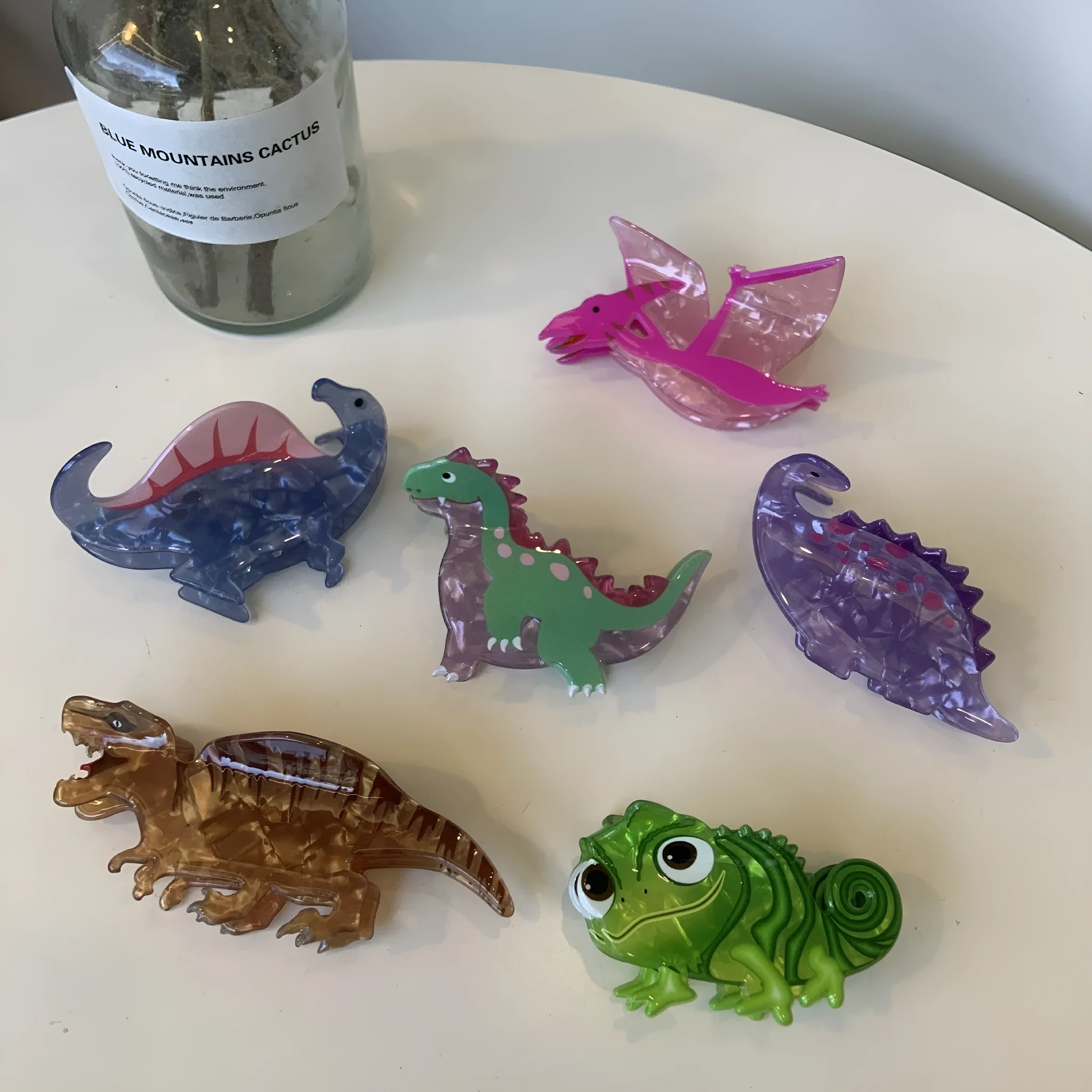 Acrylic dinosaur jaw clip hairpin showing one of six colour designs: Pink Pterosaur, Green Stegosaurus, Purple Dragon, Blue Dinosaur, Brown Tyrannosaurus, or Green Chameleon.