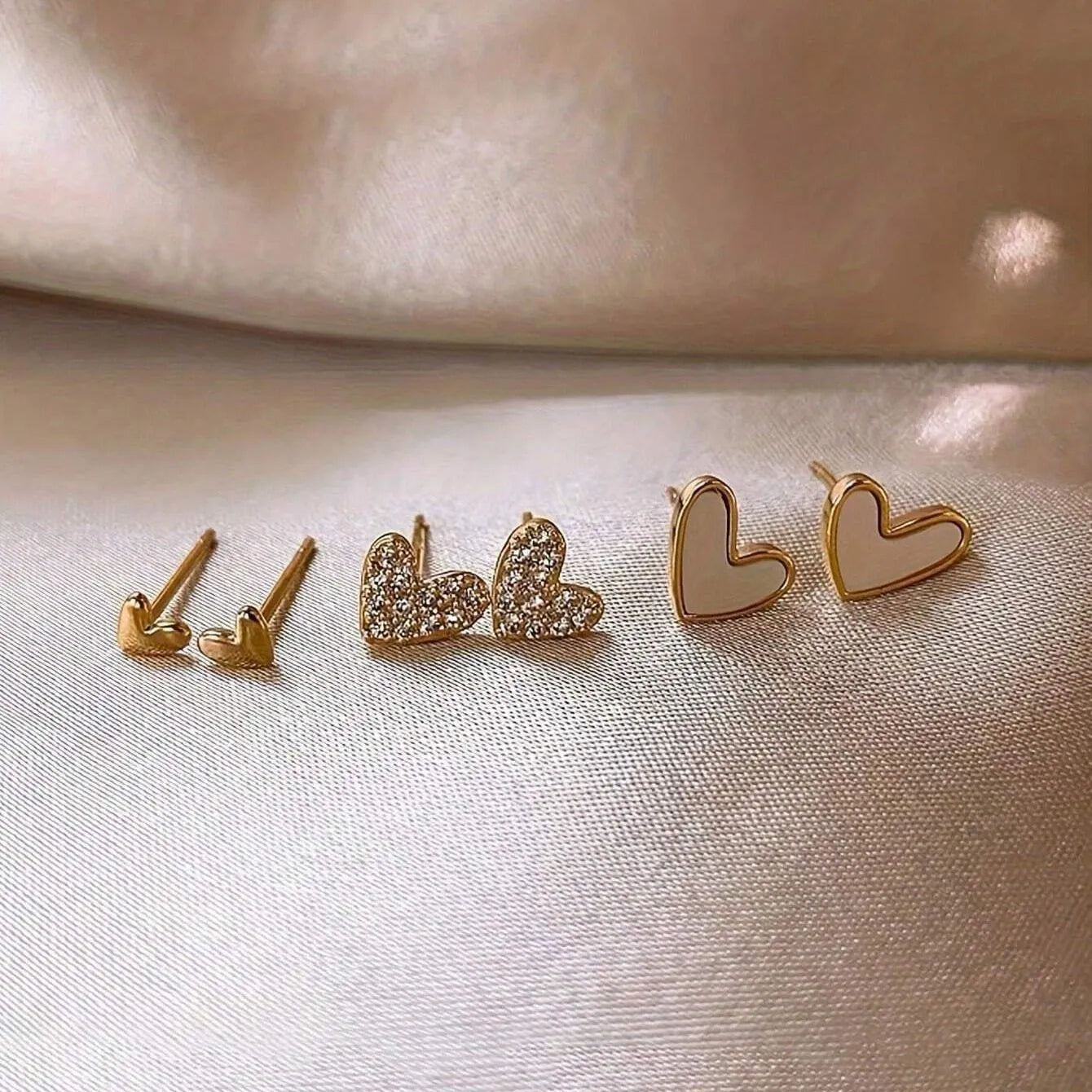 Three pairs of women's heart-shaped alloy earrings in gold, black, and pink colours on a white background.